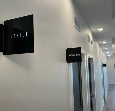 Black acrylic LED lightbox signs labeled "OFFICE" and "PACKAGE ROOM" mounted on a white hallway wall.