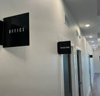 Black acrylic LED lightbox signs labeled "OFFICE" and "PACKAGE ROOM" mounted on a white hallway wall.