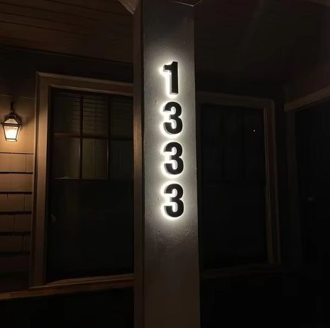 Matte black custom metal LED house numbers illuminated on a white pillar at night for clear address display.