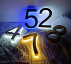 Matte black and brushed metal LED house numbers illuminated with blue, white, and warm yellow backlighting on a table.