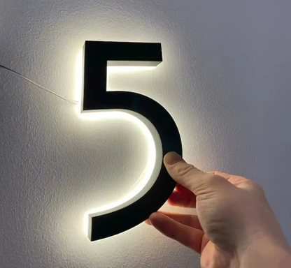 Hand holding matte black custom metal LED house number 5 glowing softly on a white wall
