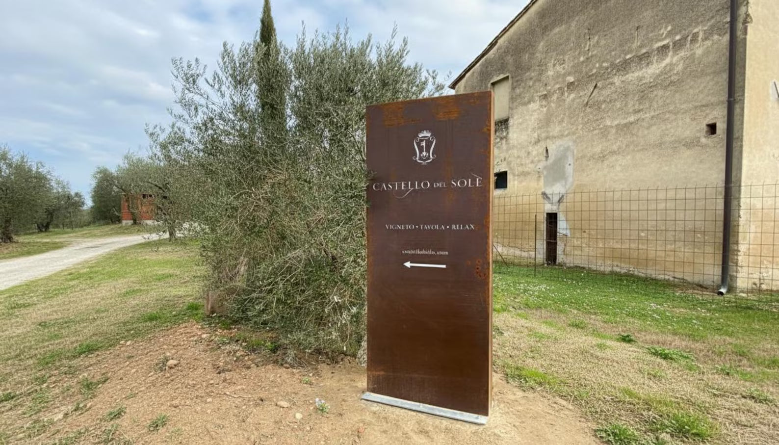Metal sign for 'Castello del Sole' in a rural setting with trees and a building.